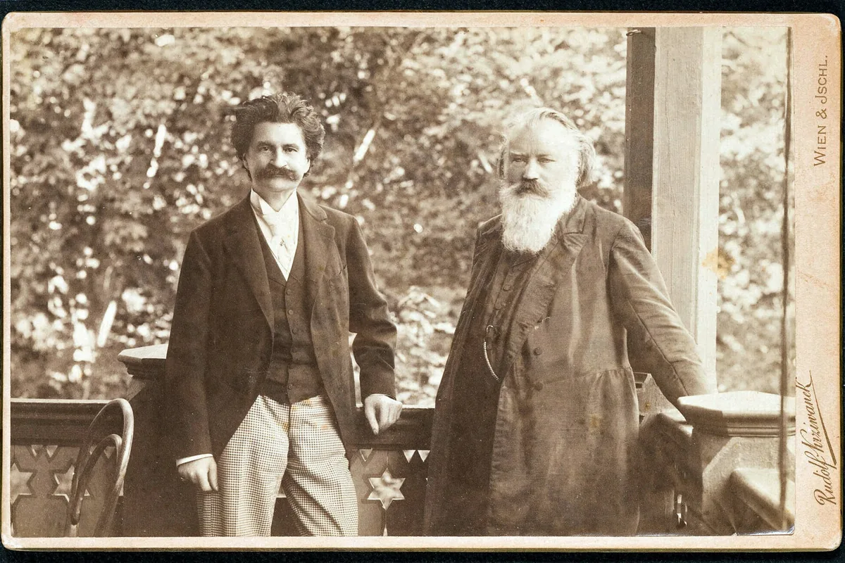 A sepia cabinet-card photograph of two men standing on a wooden veranda, one with a moustache and one with a long white beard