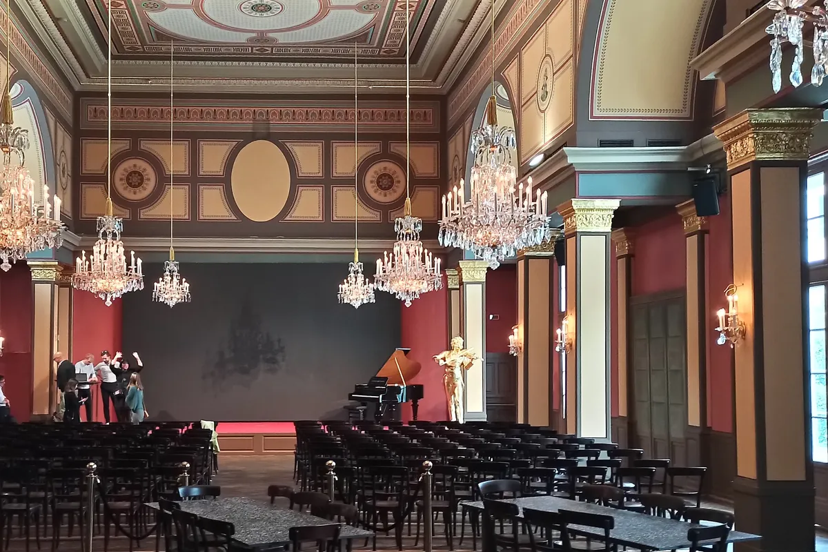 A restored nineteenth-century concert hall with crystal chandeliers under a painted ceiling, red walls with gilded capitals, a stage with a grand piano and a gilded violinist figure, and rows of dark chairs facing it