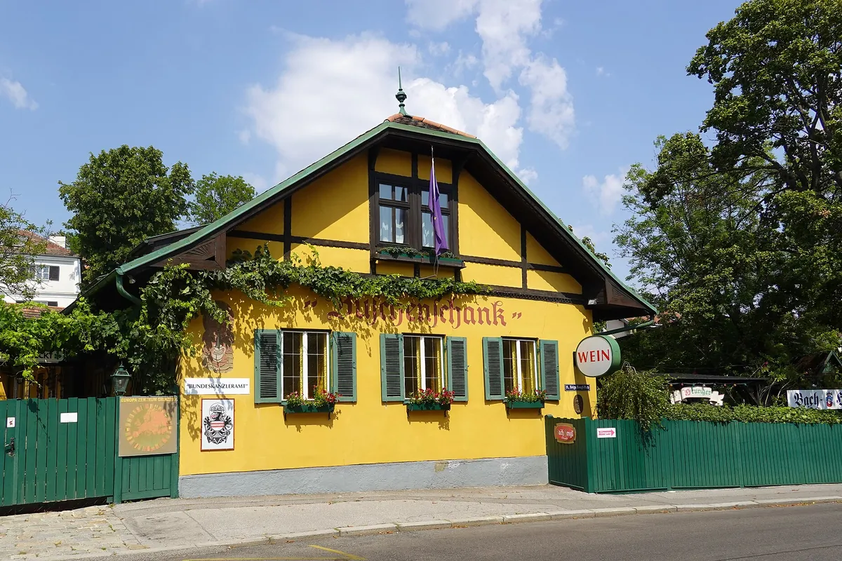 A yellow half-timbered tavern with green shutters, vines on the walls and a sign marking it as a wine tavern, beside a quiet street