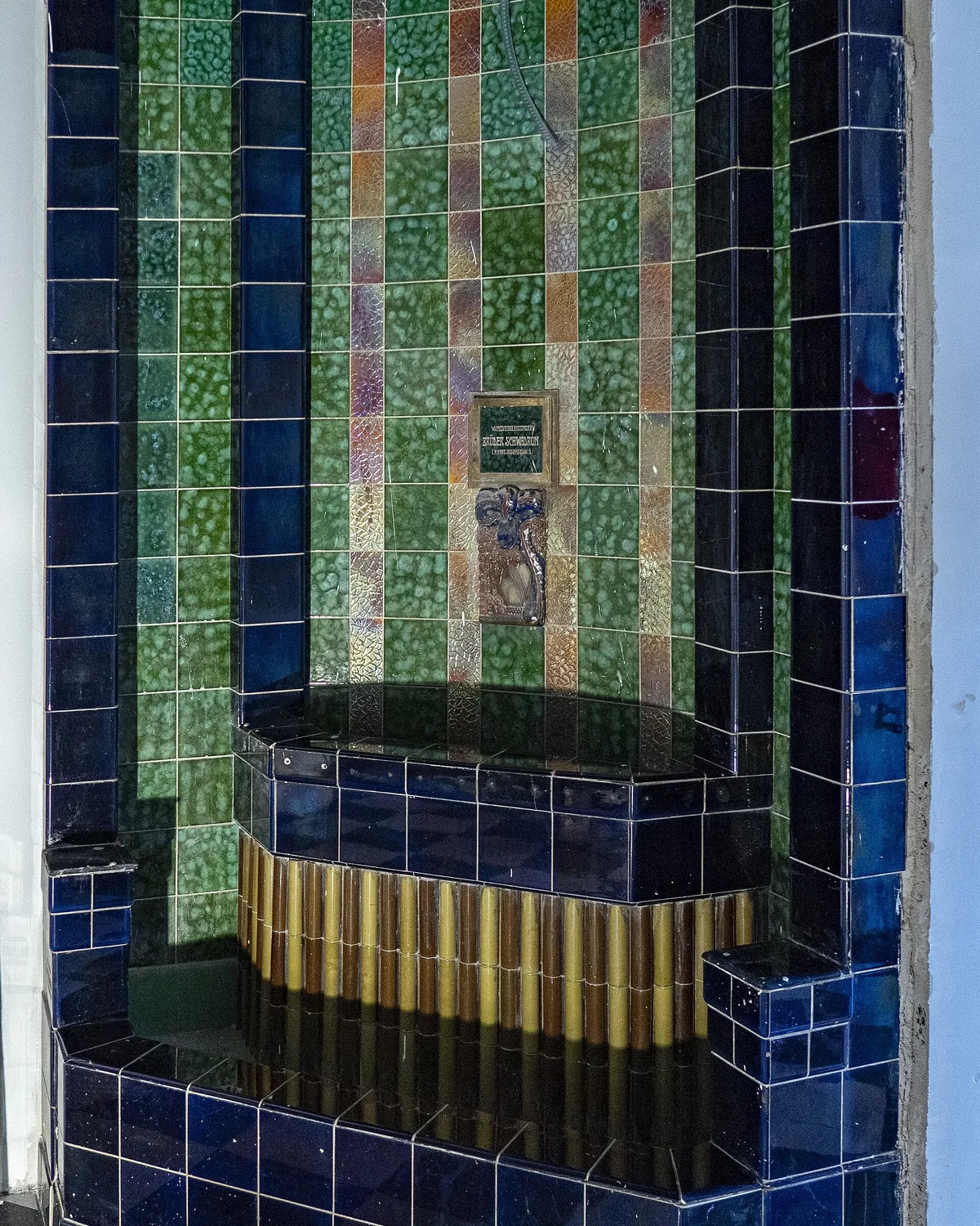 A tiled fountain niche in the foyer, lined with blue, green and iridescent tiles around a basin edged in ribbed yellow-brown tile